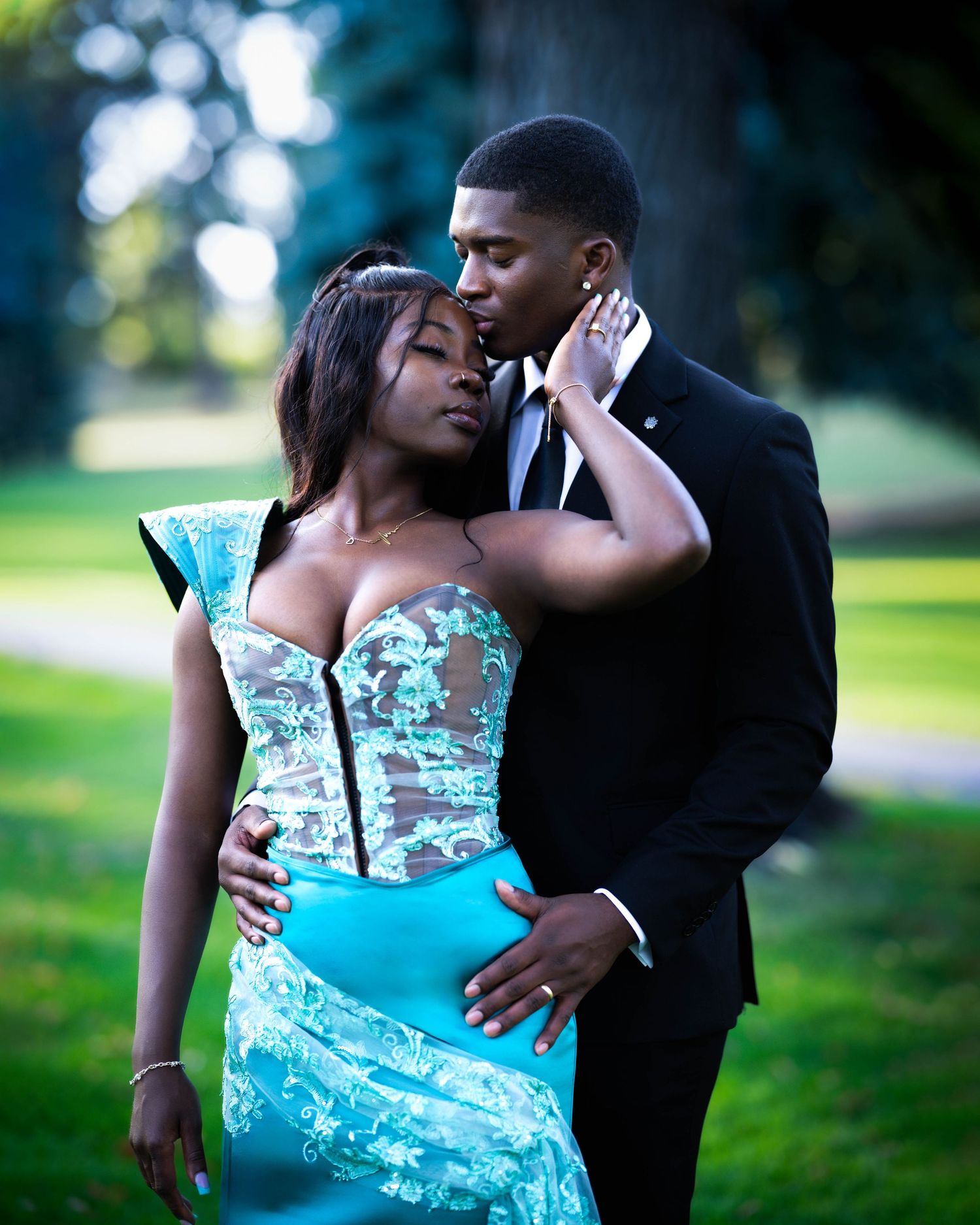 Couple in formal attire share an intimate moment outdoors, with her wearing a turquoise dress and him in a black suit.