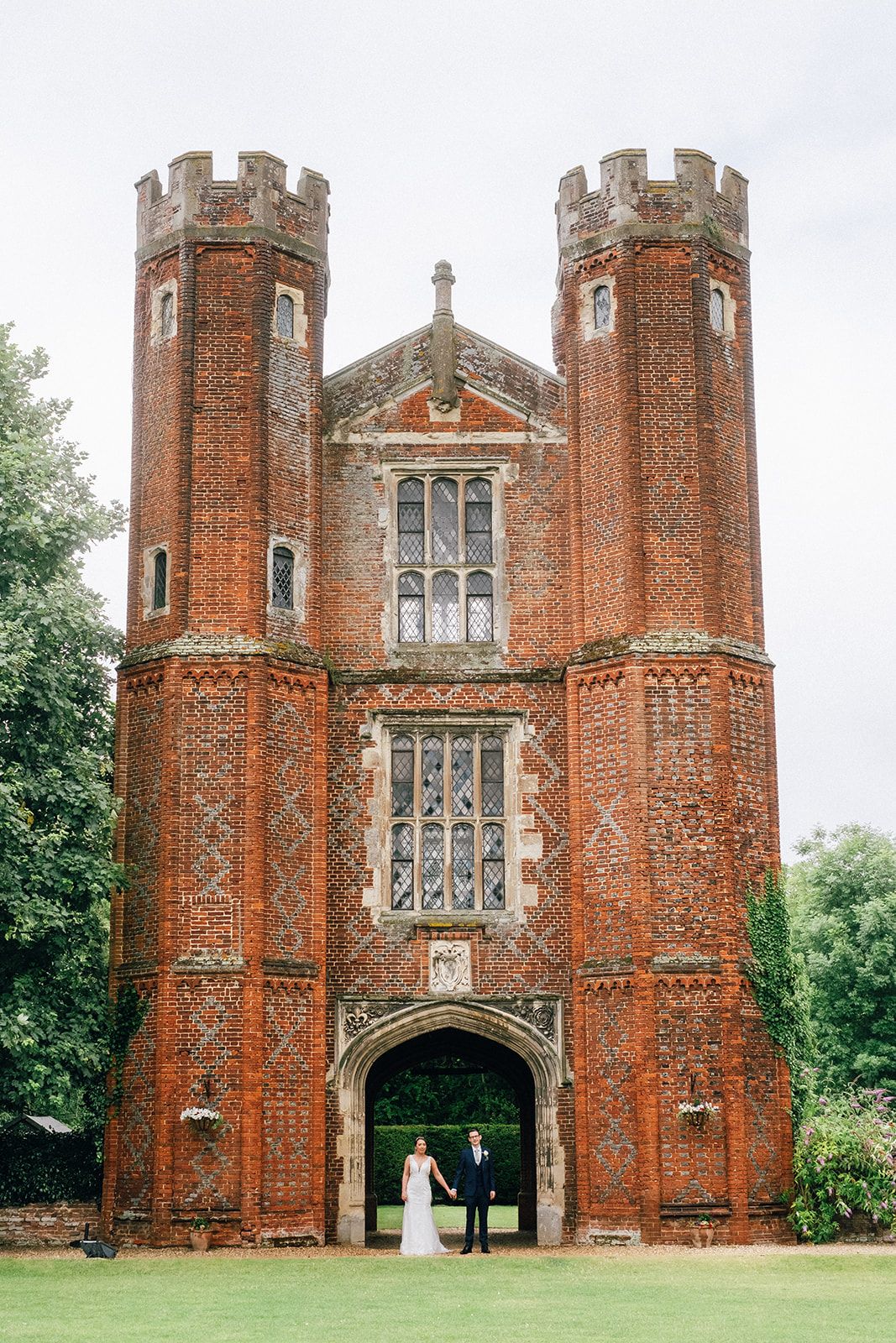 Historic red brick Tudor gatehouse with twin towers and ornate Gothic architecture stands majestically on a green lawn.