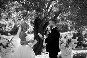 A black and white outdoor wedding ceremony under a tree with an officiant and couple exchanging vows.