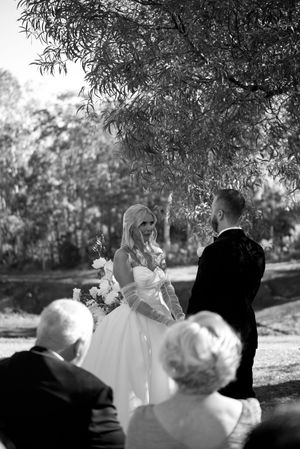 A black and white outdoor wedding ceremony under trees with guests seated and watching the couple exchange vows.