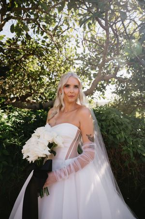 Bride in a flowing white dress poses among sunlit trees with a bouquet.