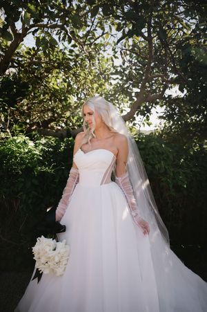 A bride in a strapless white gown and long gloves poses in a garden setting with a bouquet.