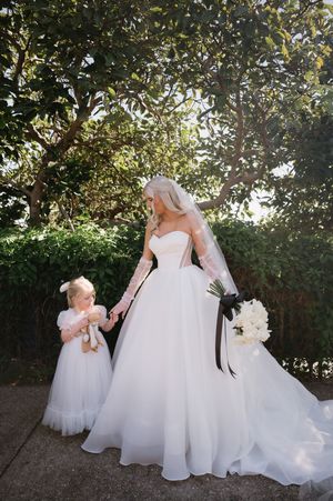 A bride in a flowing white gown poses with flower girls in matching dresses under sunlit trees.