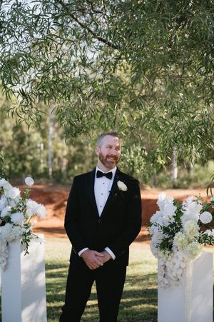 A groom in a classic black tuxedo stands between white floral arrangements under an olive tree.
