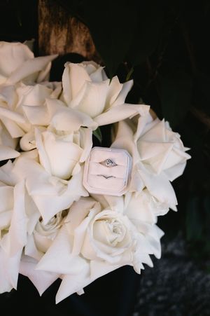 A white rose bouquet with an engagement ring box nestled among the delicate blooms against a dark background.
