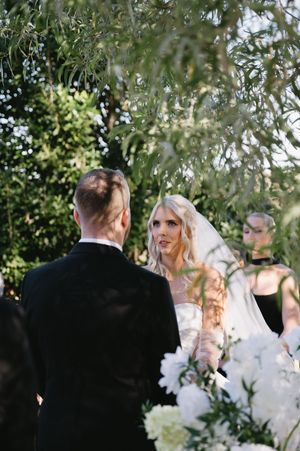 A wedding ceremony takes place outdoors under olive trees with white floral decorations.