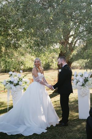 A wedding couple poses together in a romantic sequence of photos under olive trees with white floral arrangements.