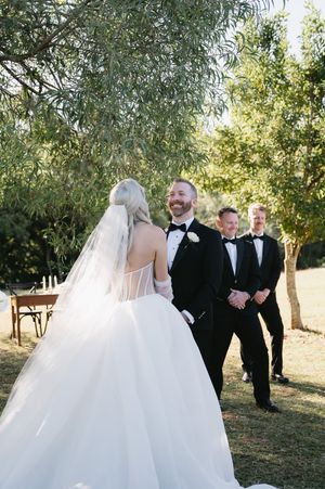 An outdoor wedding ceremony with guests in formal attire gathered under willow trees.