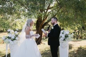 A wedding ceremony takes place under trees with white floral arrangements.