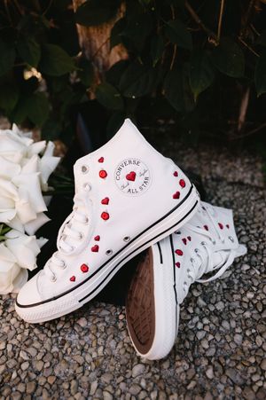 White Converse sneakers decorated with red hearts arranged on gravel with flowers nearby.