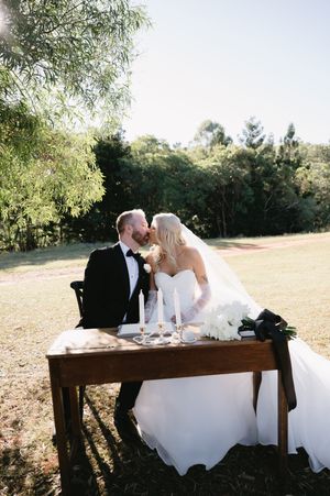 A romantic wedding couple sits together on a rustic wooden bench in a sunny outdoor setting.