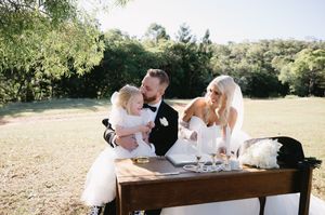 Wedding couple signing documents at rustic outdoor table during daytime ceremony.