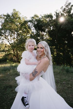 A bride in a white wedding dress holds a small child wearing a fluffy white dress and black sneakers in an outdoor setting.