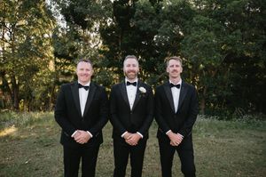 Groomsmen in black tuxedos stand together outdoors during sunset at an elegant wedding ceremony.