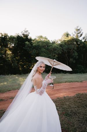 A bride in a flowing white gown holds a delicate parasol while posing in a rustic outdoor setting at sunset.