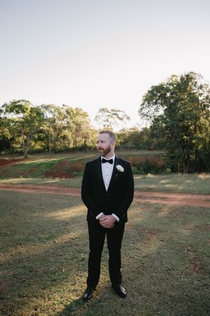 A person in a classic black tuxedo and bowtie stands on grassy terrain during golden hour.
