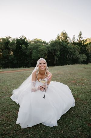 A bride in a flowing white gown sits playfully on grassy ground during an outdoor photo session at sunset.
