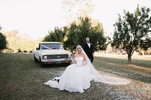 A vintage white Chevrolet car parked on grass with a wedding couple posing beside it at sunset.