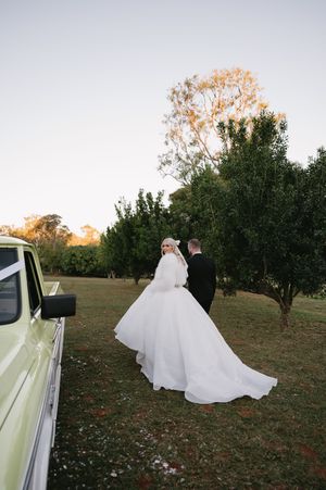 A couple in wedding attire stands in an autumn field at sunset as the bride's white dress flows in the breeze.