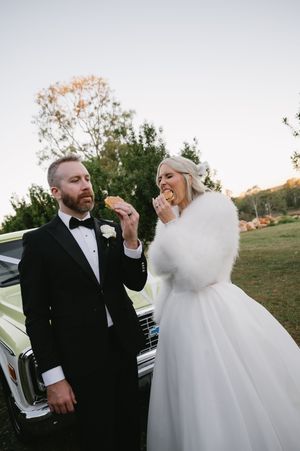 Newlyweds share a lighthearted moment next to a classic car at sunset.