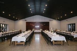 Large wedding reception hall with long banquet tables arranged in rows under string lights and vaulted ceiling.