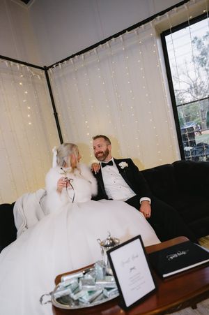 Newlyweds relaxing on couch against backdrop of twinkling fairy lights at wedding reception.