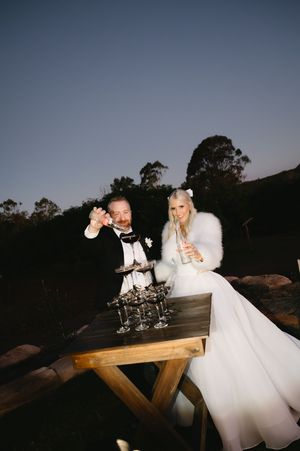 A wedding couple toasts with champagne at dusk beside a rustic wooden table with glasses.