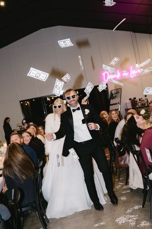 Wedding guests dance and celebrate at a reception with dollar bills floating through the air in a dimly lit venue.