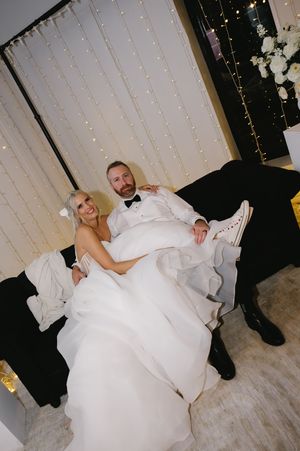 A bride and groom relax on a black couch against a backdrop of fairy lights at their wedding reception.