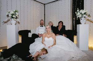 Wedding party relaxing on black couches with elegant white floral arrangements and string lights.