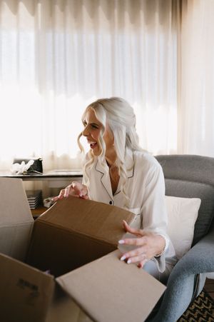 Someone in a white robe opens boxes while sitting on a grey couch during wedding preparations.