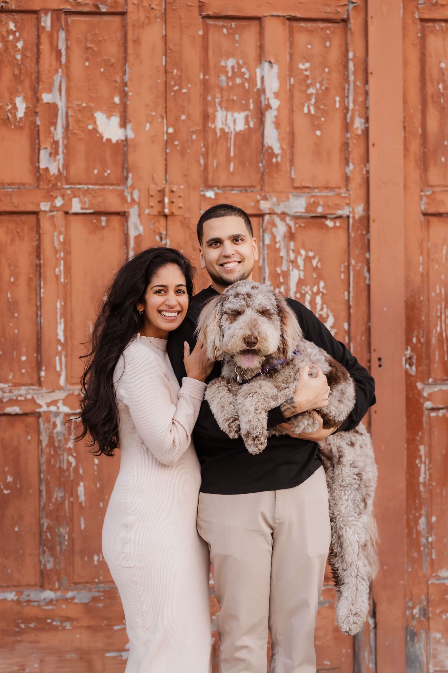 A couple in light colored outfits poses with their two fluffy dogs against a rustic orange wall.