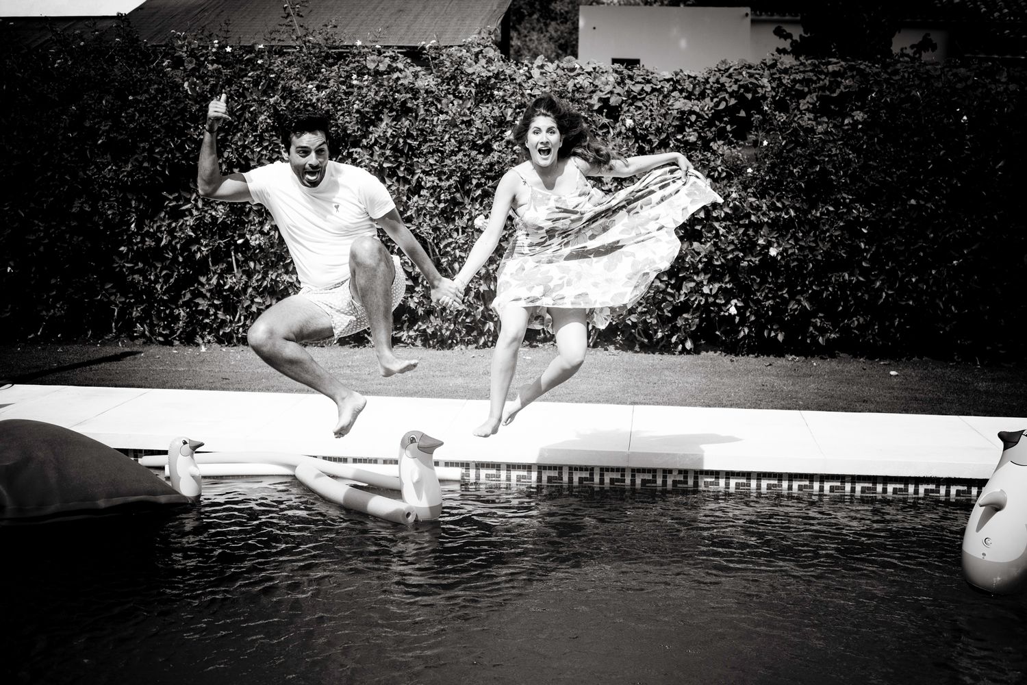 A black-and-white photo of a couple, captured jumping to a swimming pool while holding hands.