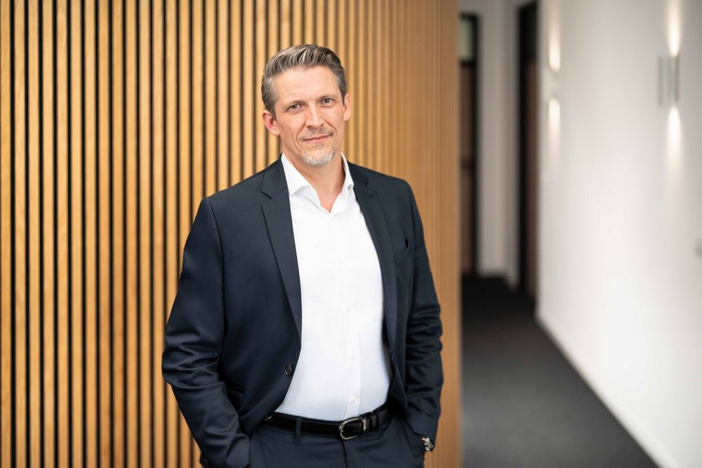 Professional wearing a dark suit and white shirt stands confidently against a wooden slat wall in an office hallway.