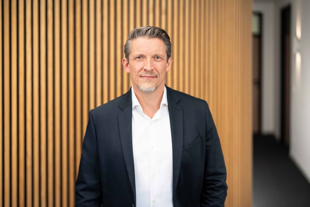 Professional headshot of person in dark suit jacket and white shirt against wood paneled wall in office hallway.