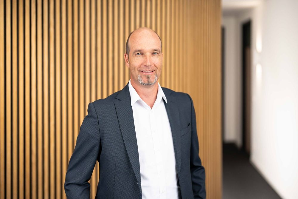 Professional in a white shirt and dark suit stands against a modern wood paneled wall in an office setting.