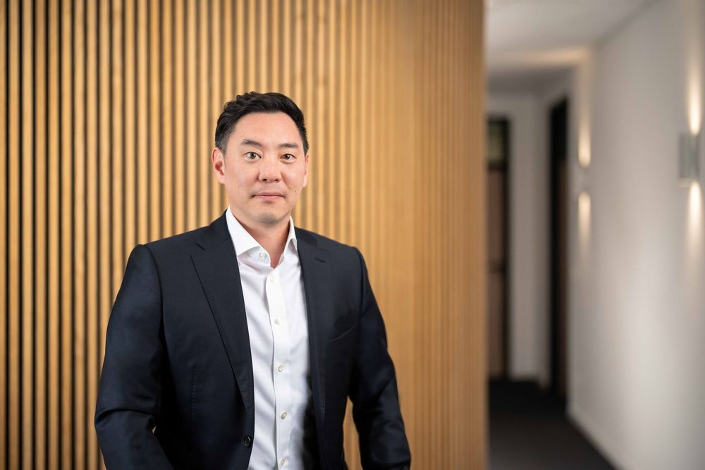 Professional headshot of someone in a dark suit and white shirt against a warm wood-paneled background.