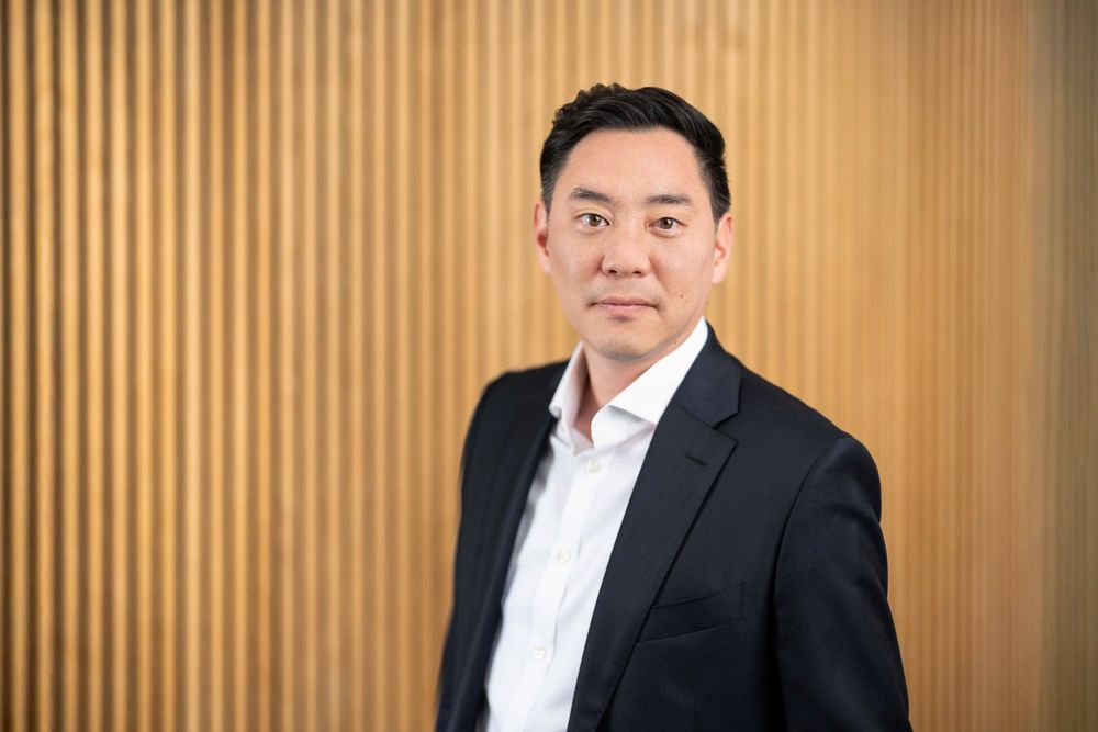 Professional business portrait against wood paneled wall with warm lighting and dark suit jacket.