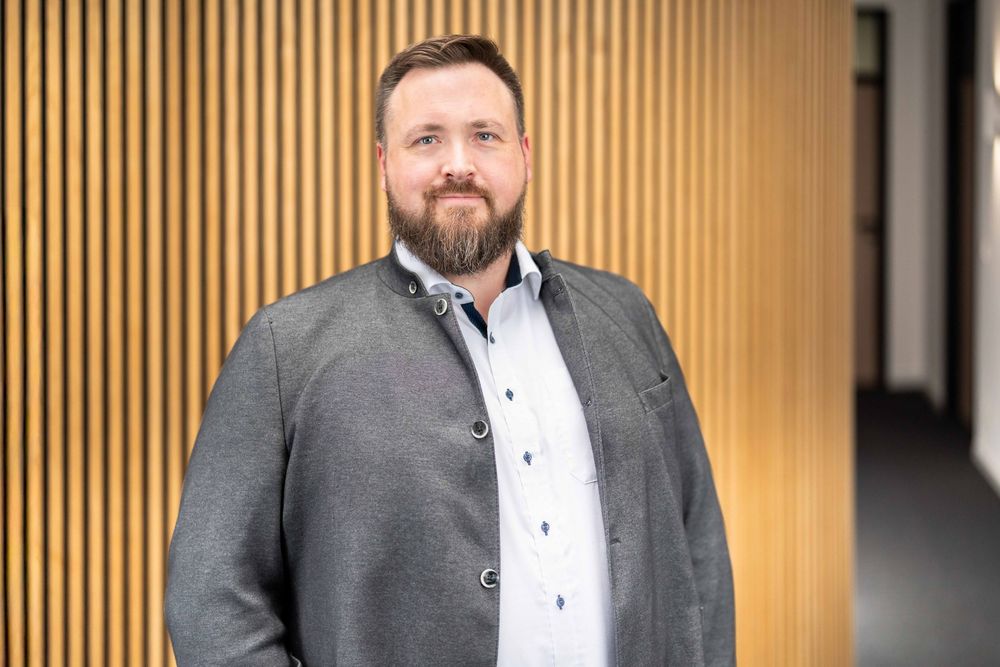Business professional in dark suit smiles while standing against wood paneled wall in corporate environment.