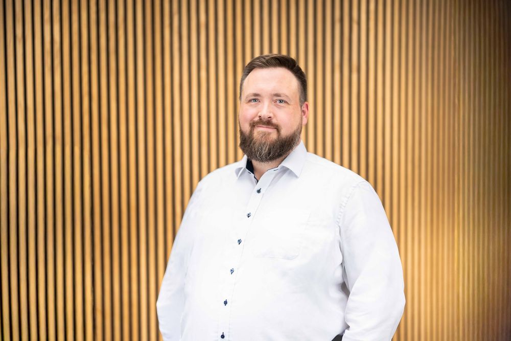 Professional portrait of person in white button-down shirt standing against wooden slat wall background.