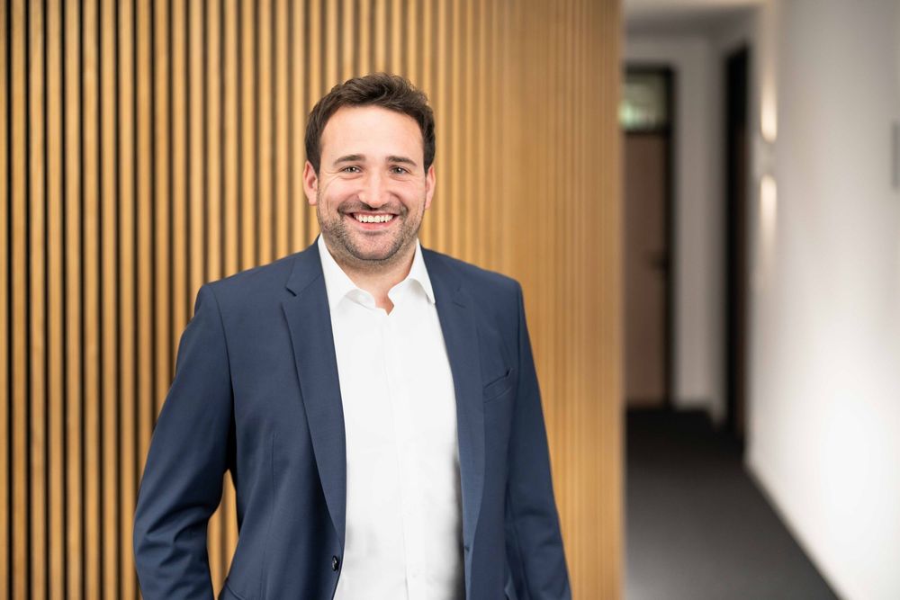 Professional portrait of person in navy suit and white shirt smiling against wood panel wall in office setting.