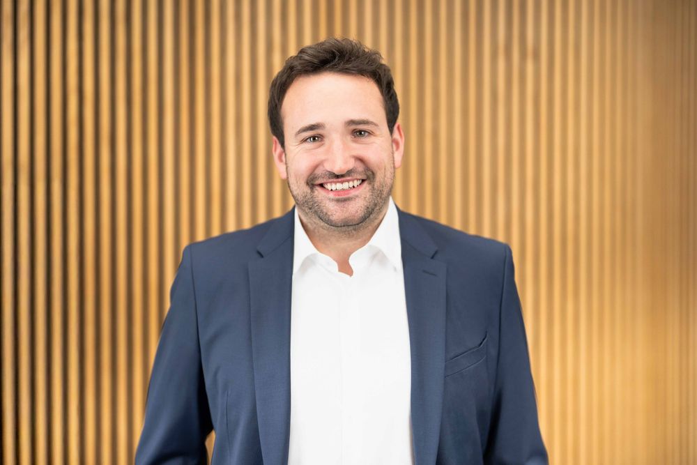 Corporate headshot of smiling professional in navy suit against modern wood panel background.