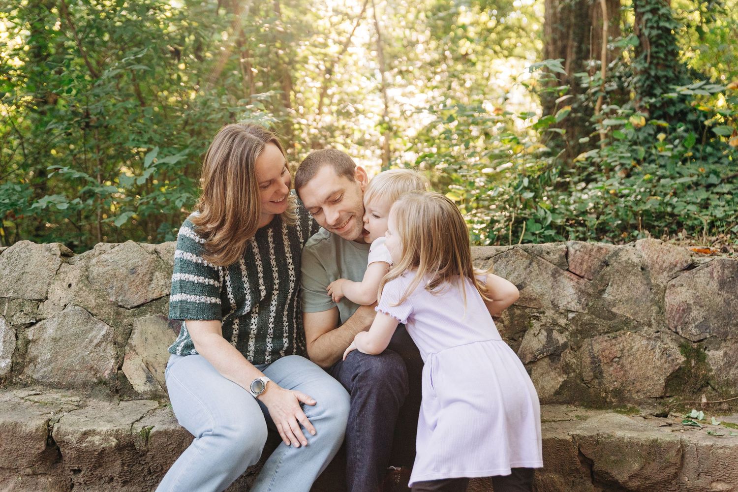 A family shares a tender moment together while sitting on stone steps in a sunlit garden setting at a Charlotte park.
