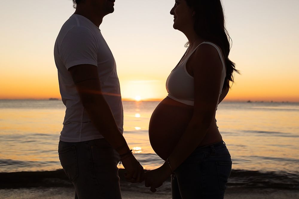 A silhouetted couple stands on a beach at sunset with an expectant mother's pregnant belly in profile.