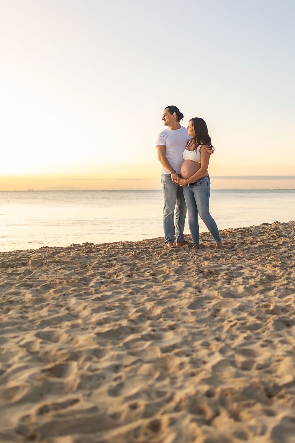 A couple stands together on a sandy beach at sunset, looking out towards the ocean horizon.