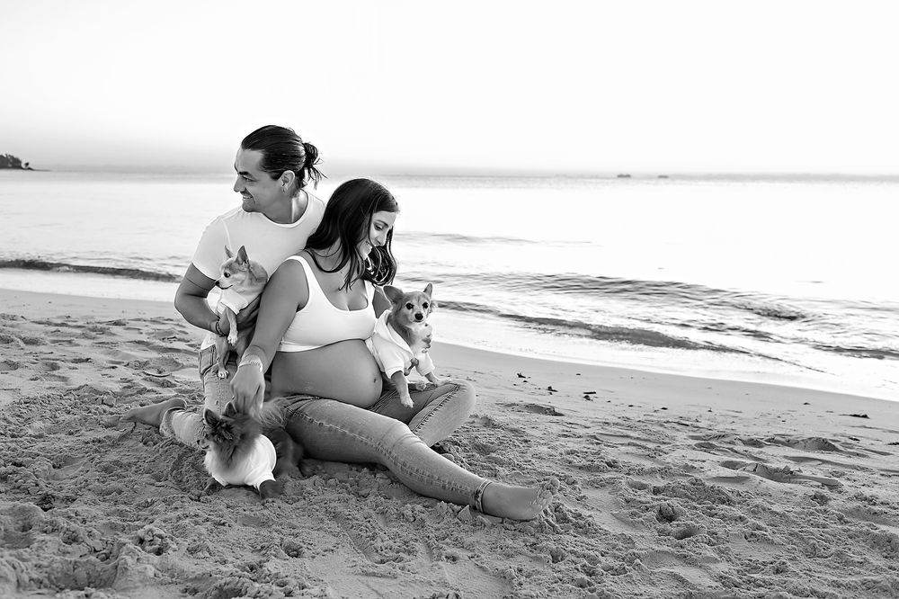 A couple sits on the beach with their dogs during a black and white maternity photoshoot.