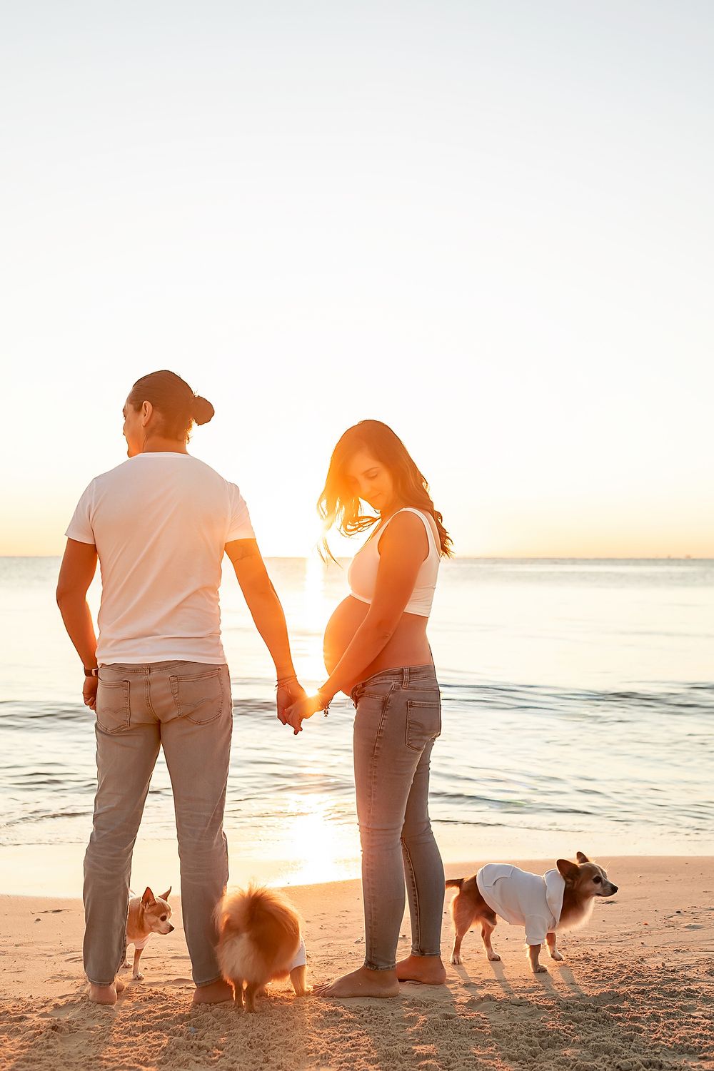 A couple and their dog stand together on a beach at sunset, silhouetted against the golden sky and ocean waves.