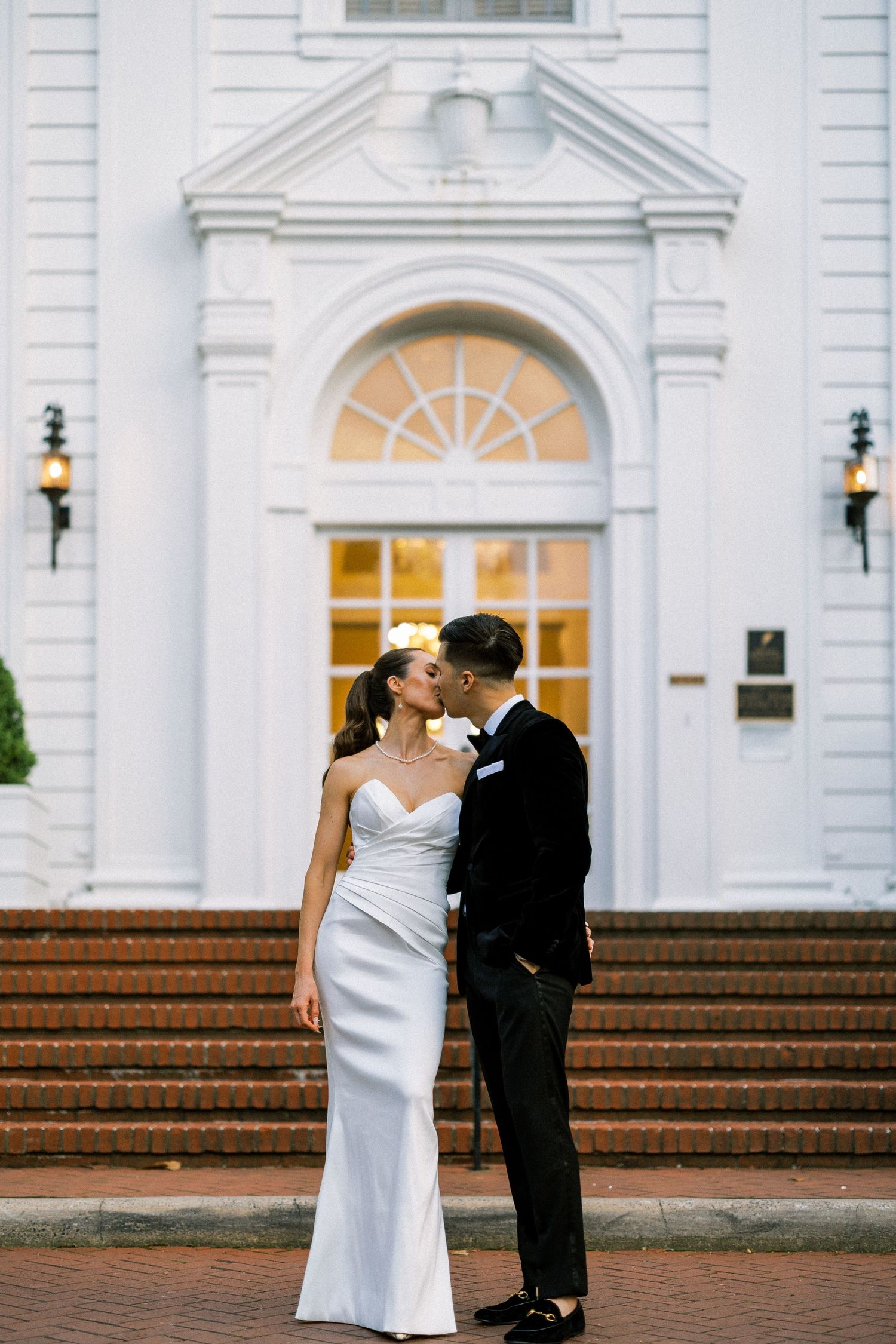 A romantic wedding couple shares tender moments on the steps of an elegant white colonial building with illuminated doorways.