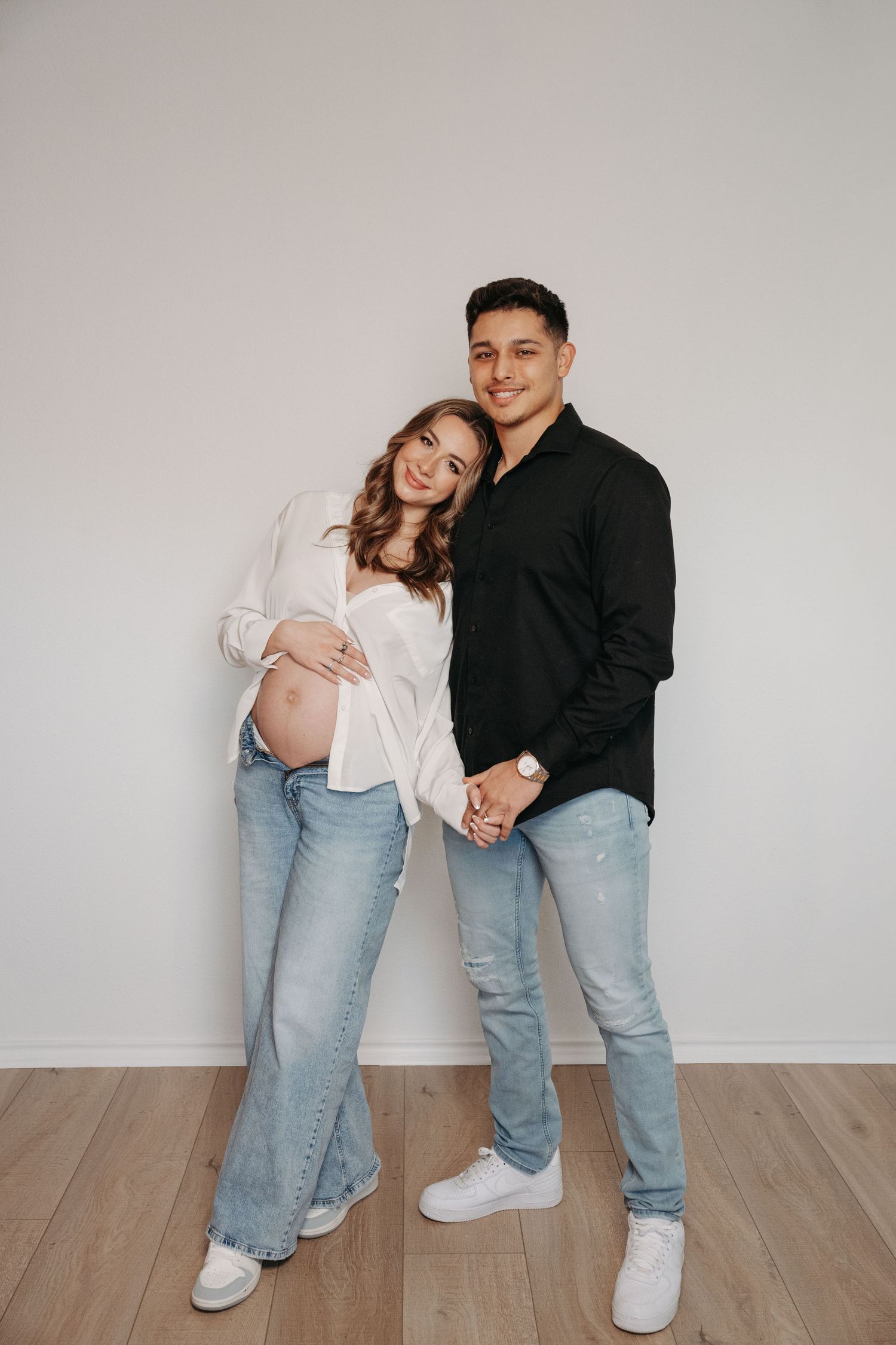 A couple in casual outfits embraces while posing against a white wall in a photo series.