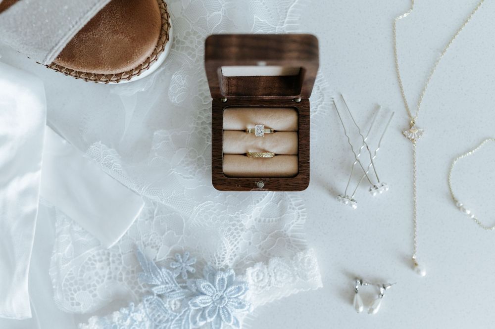 Wedding rings displayed in a wooden box alongside delicate lace and jewelry on a white surface.